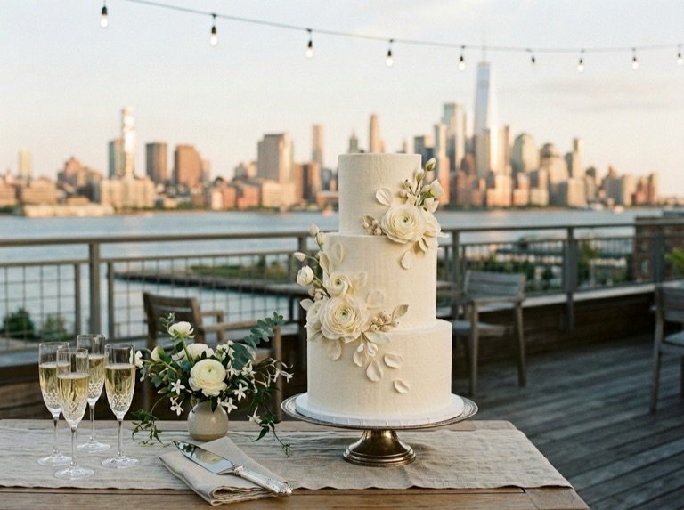The three-tier ivory cake on a silver stand, set on a rooftop table against the Manhattan skyline